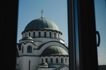 Fototapeta premium View of Saint Sava Temple in Belgrade framed by a window, with its green domes and golden cross shining under the blue sky
