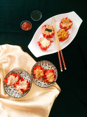 Red dimsum or Shumai or Siomay chicken and shrimp variance with topping above are served on plates,sauce,chopsticks, in black and gold background.
