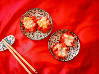 Red dimsum or Shumai or Siomay chicken and shrimp variance with topping above are served on plates,sauce,chopsticks, in red background.
