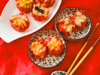 Red dimsum or Shumai or Siomay chicken and shrimp variance with topping above are served on plates,sauce,chopsticks, in red background.
