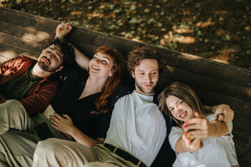 A group of four friends share a happy moment outdoors, lying together on a wooden bench under...