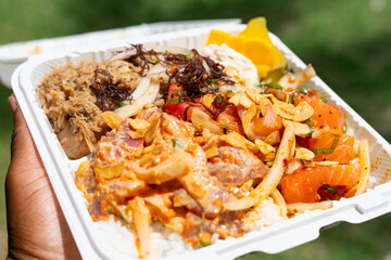 Vibrant Hawaiian poke plate with spicy ahi, salmon, and kalua pig held outdoors by a black man’s...