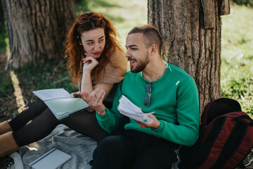 Two friends study together outside, enjoying a sunny day while sitting under a large tree.