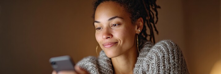 Young woman with dreadlocks wearing a knitted sweater is using a smartphone at home, smiling and enjoying mobile technology and online connectivity