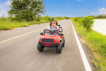 Fototapeta premium A cute little Asian girl is happily riding her toy car on a country road during her summer vacation,A little girl rides a toy car on a concrete road.