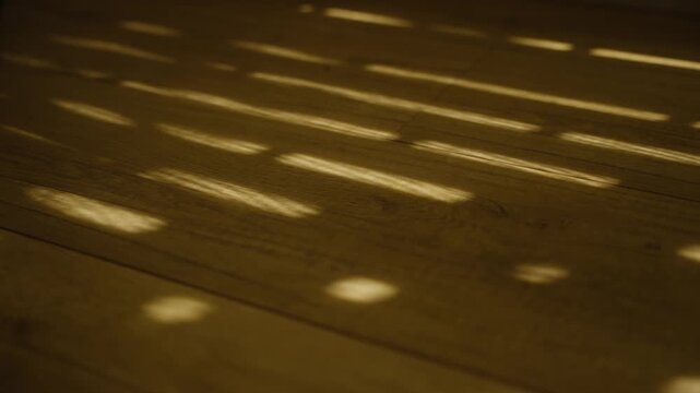 Dappled light shining through window blinds or shutters on wooden floor. Inside apartment or house warm sun is entering through cracks of mechanical or wooden shutters in early morning or afternoon