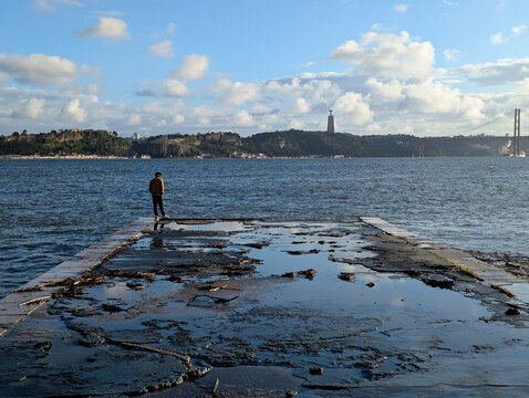 Solitary Figure on Weathered Pier Overlooking River Towards Cristo Rei Monument and 25 de Abril Bridge, Lisbon Skyline, Portugal