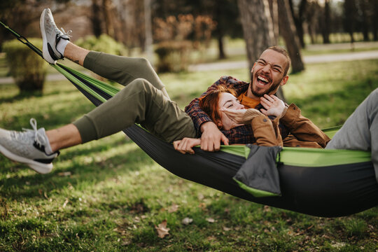 A cheerful couple enjoys a wonderful moment lying together in a hammock at a park, sharing laughter and relaxation surrounded by greenery. This scene encapsulates joy, bonding, and love in nature. - Powered by Adobe