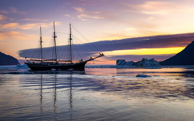 A sailing ship at anchor in Vikinge Bay, Scoresby Sund, Greenland. A warm glow in the sky is reflected in the cold arctic waters at dawn.