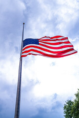An American flag flies at half mast, waving in the wind beneath a white sky with bright clouds, a...