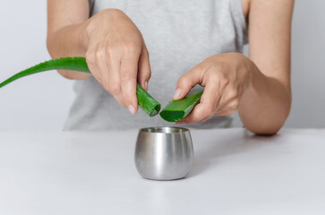 Unrecognizable woman holding two freshly cut aloe leaves above jar of gel on white table, front view