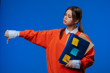 Naklejka premium A young female student gives a thumbs down gesture while holding folders and notes in her hands.
