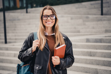 Young smiling woman holding a book outdoors wearing a stylish leather jacket during a calm day in...