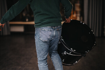 Obraz premium A person dressed casually in a sweater and jeans carries a large drum in a dimly lit music studio. The scene suggests preparation or practice, capturing a behind-the-scenes moment in music production.