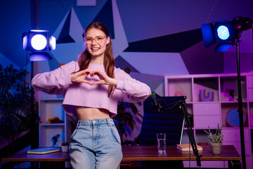 Popular female content creator in a home studio with neon lighting and microphone, making a heart...