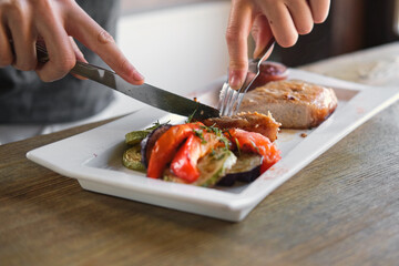 Girl cuts a cooked pork steak served on a white plate with grilled vegetables. Young woman using knife and fork to cutting meat eating in a restaurant. Balanced diet, cooking, culinary. Food concept