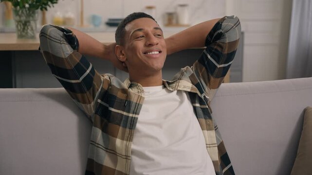 Relaxed carefree smiling African American man guy male at home relaxing in living room leaning back on couch comfortable sofa enjoying new home own tenant weekend indoors break pause thinking dream