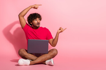 Young stylish man with curly hair sitting cross-legged holding a laptop on a pink background and...