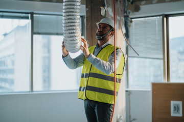 Professional engineer in a safety vest and helmet examining ventilation duct work during a building...
