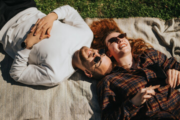 A cheerful couple lays on a blanket outdoors, basking in the sunlight, enjoying their time...
