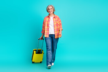 Joyful mature woman with yellow suitcase on teal background representing leisure travel and casual...