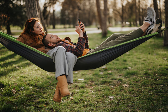 A loving couple spends time in a park, lying together in a hammock while savoring a calm moment surrounded by nature and vibrant greenery.