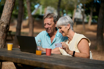 Senior couple making online purchases in park using laptop and credit card