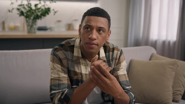 Pensive serious African American man at couch considering problem solution solving difficult trouble thoughtful hesitate male guy thinking rubbing hands think in apartment brainstorming challenge