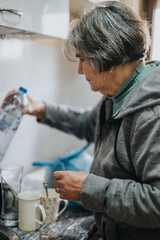 Elderly woman preparing a drink in a home kitchen with glass and mugs visible on the countertop.