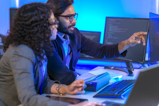 A senior software developer or team lead points at code on a screen, mentoring a female colleague. Diverse professionals collaborating on a project in a modern IT office.