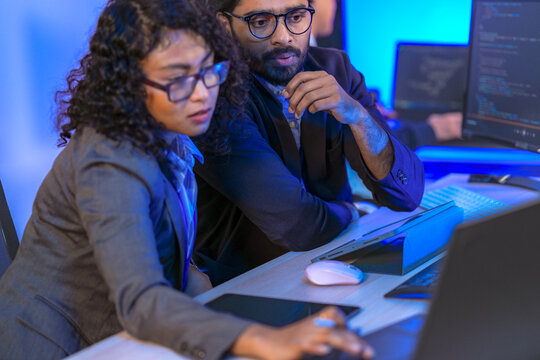 A senior software developer or team lead points at code on a screen, mentoring a female colleague. Diverse professionals collaborating on a project in a modern IT office.