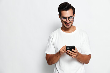 Fototapeta premium Smiling young man wearing glasses and a white t-shirt, happily interacting with his smartphone, standing against a solid light background. People lifestyle concept.