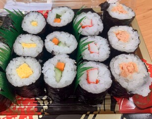 A neatly arranged set of twelve sushi rolls a colorful mixed ingredients like crab, egg, cucumber, tuna, wrapped in seaweed and accented with fresh green leaves serving in black vintage plate