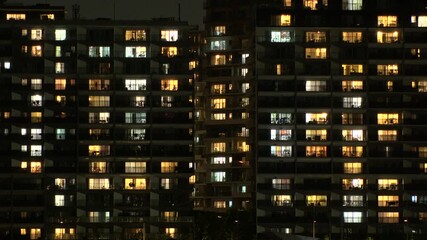 TOKYO, JAPAN - AUG 2025 : Close up shot of illuminated modern high rise residential apartment building with glowing windows in dense urban cityscape at night. Lifestyle and real estate concept video.