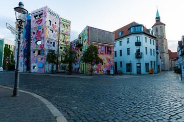 A brightly painted building with playful shapes and colors stands out in the street view. Cobblestones and surrounding houses complement the scene in morning light.