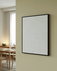Blank square canvas framed with a thin black border mounted on a textured wall in a home interior next to a blurred dining table setting