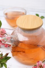 Tasty rose tea in glass teapot, flowers and leaves on white wooden table, closeup