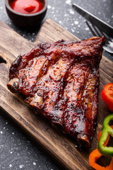 BBQ pork ribs from pork tenderloin with vegetables, ketchup on board and dark background close up vertical photo