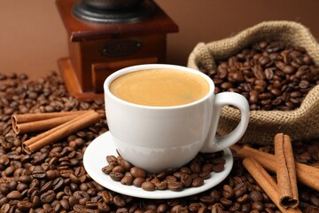 Cup of hot coffee, beans, grinder and cinnamon on brown background, closeup