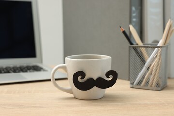 Happy Father's Day. Cup with paper moustache and laptop on wooden table, closeup