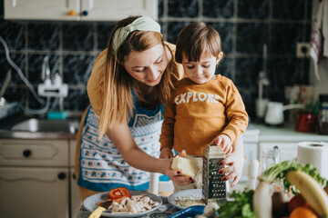 A cheerful mother guides her involved young child in preparing a meal with various fresh...