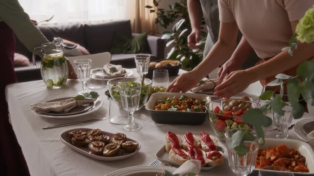 Family members setting dining table with cutlery and various dishes while preparing for festive dinner in living room