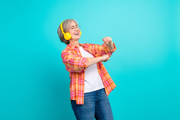 Elderly woman in a checkered shirt enjoying music with headphones against a turquoise background, exuding joy and energy
