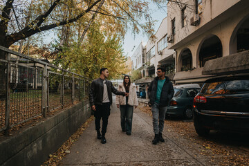 Obraz premium Three friends walking together through a city street during autumn, surrounded by falling leaves.