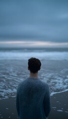 Fototapeta premium A lone man in a wool sweater stands by the ocean at twilight