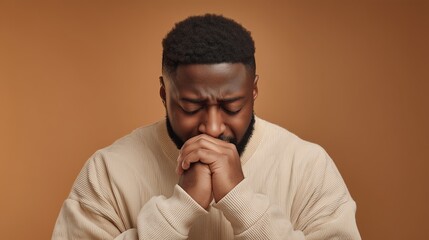 Obraz premium A young African man with short black hair is praying with his hands clasped together. He wears a light-colored sweater against a warm brown background.