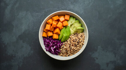 Overhead view of a healthy and colorful bowl containing cubed sweet potatoes, sliced avocado, shredded red cabbage, and quinoa on a dark textured surface.