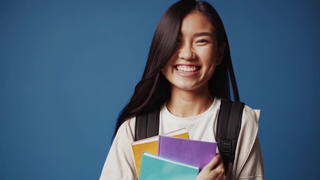 Education and students. Happy asian woman, holding notebooks and laughing, smiling at camera, enjoys going to University or College, blue background
