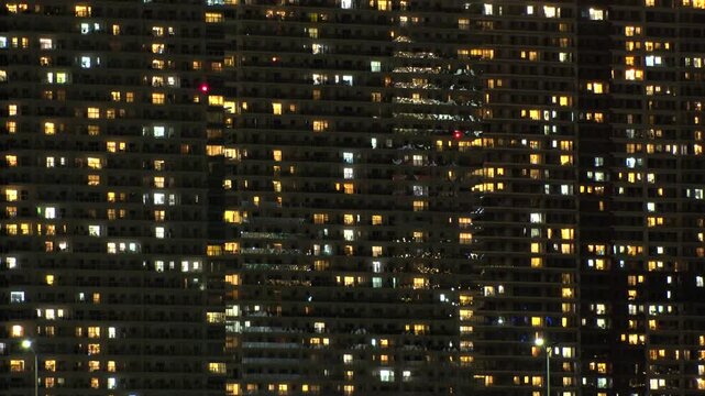 Time lapse shot of illuminated modern high rise residential apartment building with glowing windows in dense urban cityscape at night. Lifestyle and real estate concept.