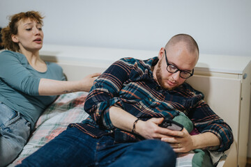 A couple is seen in a bedroom, with the man distracted by his phone and the woman trying to engage him, showcasing themes of disconnected relationships and modern communication challenges.